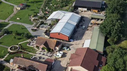 Ant Saddington/BBC An aerial drone shot of the whole site showing several medium and large buildings or farm sheds, with three police cars parked in the courtyard. There are also formal gardens on the left side and lots of greenery.