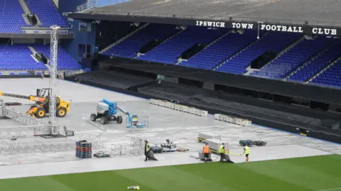 Workers wearing hi-vis jackets walking on the pitch at Portman Road. The pitch is mostly covered in protective plastic boards, but part of the playing surface remains exposed. There is a yellow heavy machinery vehicle putting large metal structures in place where the boxing ring will be. A stand with blue seats is on the right of the picture.