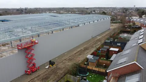 A drone shot of the incomplete warehouse - a grey structure that stretches across the frame several metres in front of a housing estate. The estate backs directly onto the warehouse land.