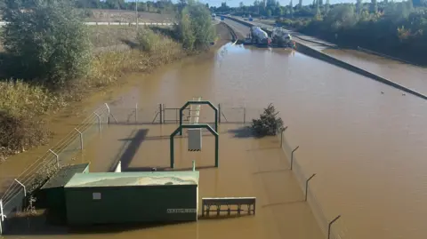 Tony Fisher/BBC A road and its pumping station submerged under floodwater with tankers in the background pumping out the water. 