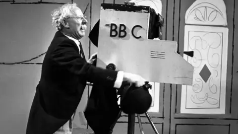 A black-and-white mid-shot of a man in his 50s, made up to look older with white hair and a white moustache. He is wearing a black tailcoat, a white shirt and grey trousers, and is pictured side-on from his right-hand-side. He is holding a black bowler hat in his right hand, which is stretched out alongside a mock-up of a 1930s-style BBC television camera which he is pretending to operate. Behind the camera is an impressionistic scenery flat depicting two ornate windows.