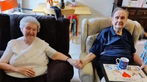 Fiona Davidge Photograph of Avril (left) and Clifford (right) sat on two armchairs. They hold each other's hands in the centre of the image and smile at the camera. Avril wears a white t-shirt, while Clifford wears a navy polo shirt. Clifford has a tray with a cup on it.