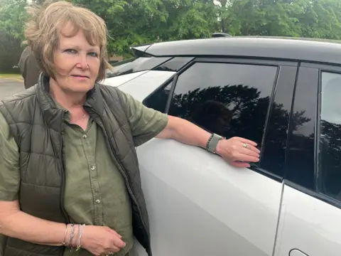 Chrissanne Robertson standing next to her damaged car. She is wearing a green shirt and a black padded gilet. Her arm is next to the window seal at the rear of her silver car