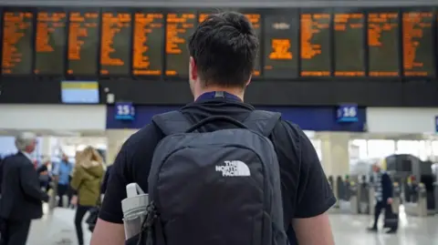 Man stood in front of train boards at Waterloo station.