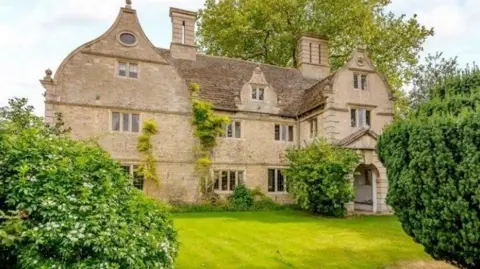 Historic house in beige stone- with green lawn in front of it and trees on either side.