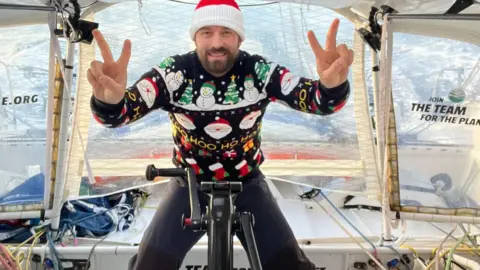 Sam Goodchild A man in a Christmas jumper and hat gives two V for victory signs in the cockpit of a yacht.