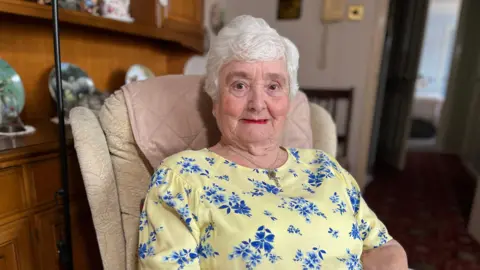 BBC An older lady, dressed in yellow, smiles at the camera