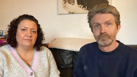 A woman with dark, curly hair wearing a white cardigan and a man with swept back brown hair wearing a blue sweatshirt sit on a sofa together.