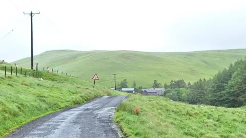 Laura Day It is a wet day and a wet single-track tarmac road with grass on either side stretches along towards a farm. There are electricity poles stretching down towards it and beyond is a grassy, green hill.