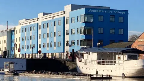 A photo of the Gloucestershire College campus next to Gloucester Docks. The building has blue and white cladding and is next to canal basin, where a white boat is moored up
