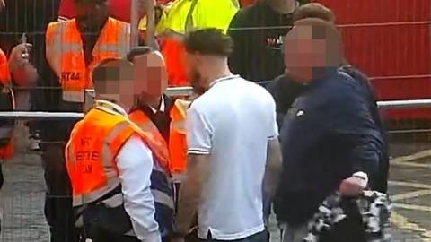 A CCTV image of a man wearing an England Tshirt with his back facing the lens, as he is surrounded by event stewards. 