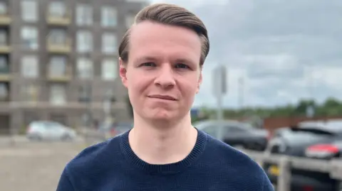 Joshua Reynolds stands wearing a blue jumper. He has combed brown hair. Behind him in soft focus is a grey building and a parked car can be seen over his left shoulder. 