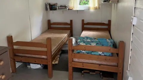 Two single beds in the bungalow's bedroom, a bookshelf is on the wall behind them with books on 