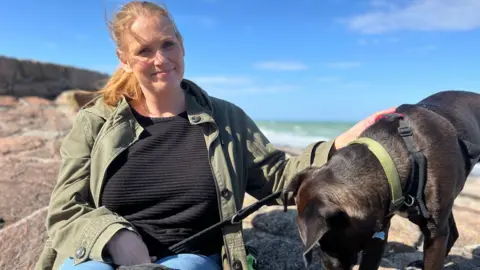 Candice Wilson is a woman with strawberry-blonde hair, wearing a black jumper and green coat. She is smiling into the camera and is sitting on a rock by the beach. She has her black dog, Theo, to her right. He is looking down at the rock.