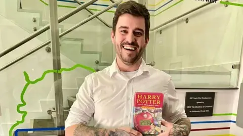 Daniel Pearce standing in front of a glass panel holding the Harry Potter book with both hands. 