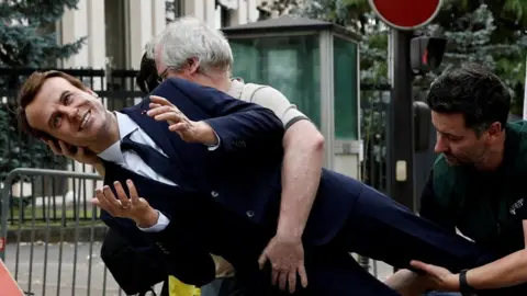 Two men carry the suited waxwork of Emmanuel Macron outside the Russian embassy in Paris