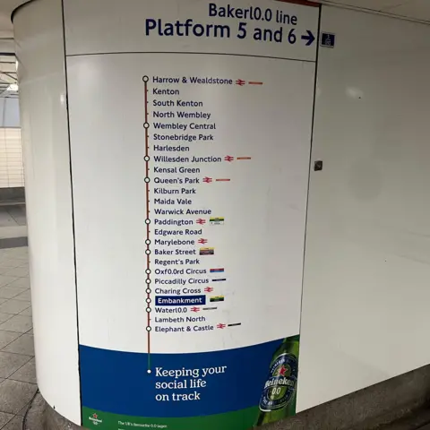 A tiled underground corridor wall showing a vertical Bakerl0.0 line route map labelled “Platform 5 and 6”, with station names listed from Harrow & Wealdstone to Elephant & Castle. An advertising panel is positioned below the map.