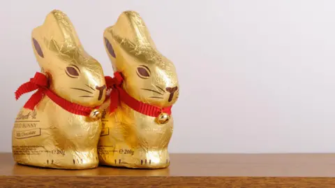 Two Lindt Easter chocolate rabbits next to each other on a wooden table with a blank beige wall behind. The rabbits are wrapped in gold coloured foil and have a red ribbon around their necks with a bell on.