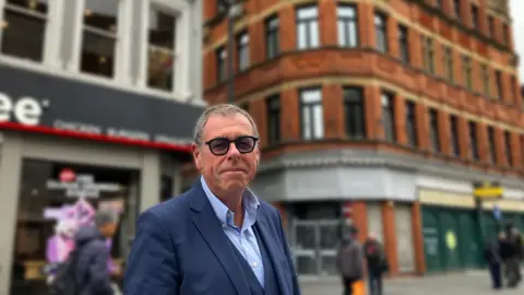 Ian Silverberg, who is middle-aged, wearing a blue suit and has short hair and glasses, stands in Whitechapel in Liverpool in front of a lamppost fitted with a speaker that plays classical music to deter youths