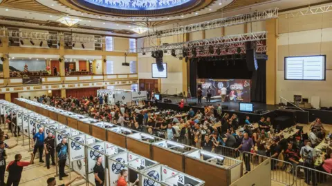 East Riding of Yorkshire Council Interior of Bridlington Spa with rows of partitioned dart boards in the foreground in front of rows of desks with dozens of people sitting at them and a raised stage in the background