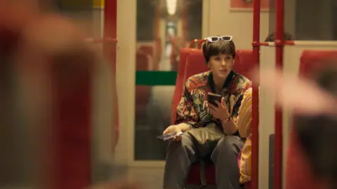 In a still from the Nightsleeper drama, the actor Alexandra Roach is seen sitting in a seat on a train. She has dark glasses on her head and is holding a mobile phone in each hand. The shot is taken down the carriage's corridor and red seats, most of them empty can be seen.