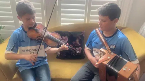 Two young boys are sitting on a yellow sofa. They are wearing light blue and white jersey-style tops. On the left the boy is playing a fiddle and the boy on the right is playing an accordion. 