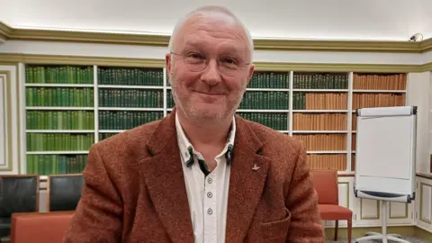 Prof Rob Ó Maolalaigh is wearing a rusty red coloured tweed jacket and a white shirt. He is smiling as he sits in the national library.