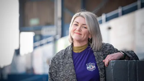 Harry Atkinson/Plain Creative Close-up of Emma Thompson at Workington Town's stadium. She has shoulder-length blonde hair and is wearing a grey coat. She is smiling as she looks into the camera.