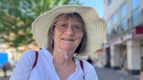 Claire Salaman is wearing a wide‑brimmed sun hat and glasses and is standing on a sunlit street between buildings.
