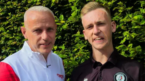 Casey Young A man wearing a white top is stood shoulder to shoulder with a younger man, wearing a black polo.