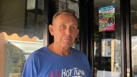 Chris Mount looking at the camera and stood in front of his shop. He is wearing a blue t-shirt. 