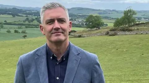 David Smith MP is smiling. He has short grey hair and is wearing a light blue blazer and dark blue, open-necked shirt. He is standing in a field with more rolling countryside, trees and dry-stone walling in the background.