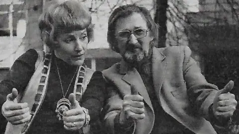 Northern Theatre A black-and-white photo of a woman and a man sitting next to each other with their thumbs up. The man has long hair and a beard and wears a jacket and glasses. He is looking straight ahead. The woman has short hair and wears a dark top, sleeveless jacket and large medallion. She is looking towards the man.