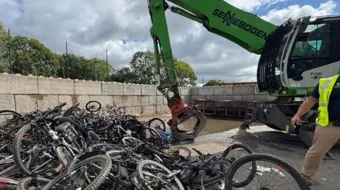 A large pile of bikes, with breezeblock walls surrounding them. A green digger with a grapple attached can be seen to the right of the image, waiting to grab and move the bikes. 
