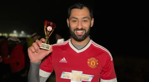 Ismail smiles at the camera as he holds the trophy after the race. He's wearing a Manchester United football top with a Jersey Spartan AC race number on it. He as a grey lycra top under his shirt. He has a short dark beard and short dark hair. Runners are standing behind him in the dark.