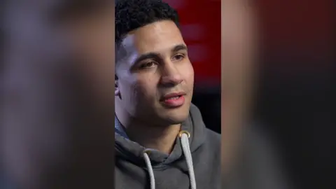 Delicious Orie looks off camera. He has short black hair and is wearing a grey hoodie with white drawstrings.