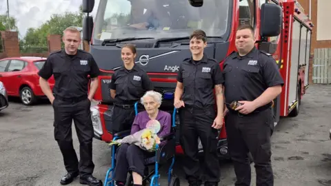 Roxburgh House An elderly woman in a wheelchair sat in the middle of four firefighters who are stood infront of a fire truck