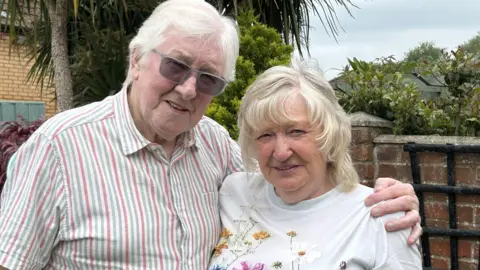 A couple with white hair stand with arms round each other in a garden. He is wearing a striped button-up shirt and sunglasses. She is wearing a top with a flower design on it. Behind them, a brick garden wall and plants are visible. 