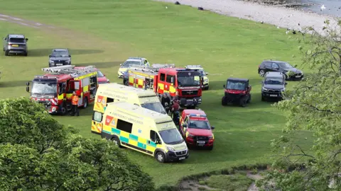 Ambulances, fire engines and other vehicles parked on a trimmed field close to a river meandering nearby