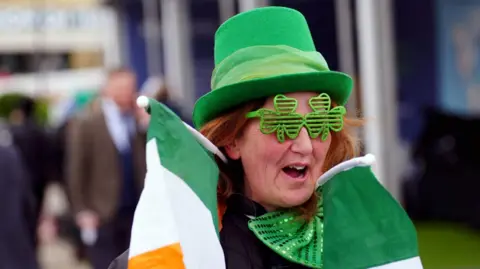 Mike Egerton/PA A woman smiling and wearing a green hat and glittery green four-leafed clover glasses. She has red hair and is carrying two Irish flags around her neck.