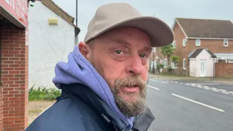 Andrew Turner/BBC Ade Evans is bearded, and is wearing a blue jacket and light blue hoodie and a beige baseball cap. He is standing outside the Chinese take-away on Acle High Street.