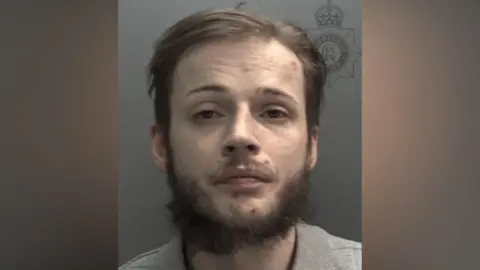 A police head and shoulders shot of a man with brown hair and a brown beard looking into the camera with a serious expression and frowning. He is wearing a grey top and the background is grey.