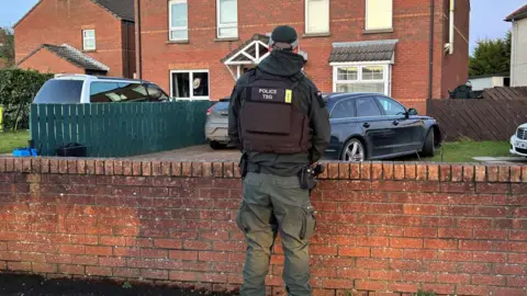 A police officer wearing a flack jacket is standing looking toward a red brick house. The officer is in full uniform which is dark green.