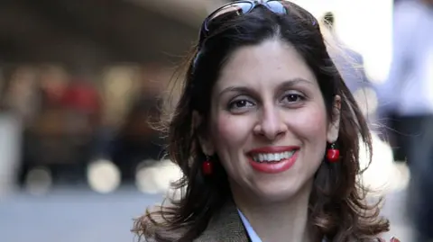 HANDOUT A family handout file photo of Nazanin Zaghari-Ratcliffe, as the British-Iranian charity worker. Nazanin has sunglasses on her head and has red earrings dangling from her ears as she looks towards the camera smiling. The background behind her shoulders is blurred.