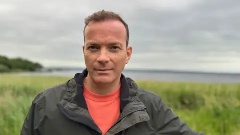 Simon Chambers. He has short brown hair, wearing a khaki raincoat and orange t-shirt. He is standing on the shores of Lough Neagh.