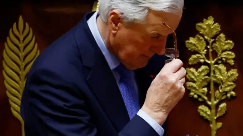 Reuters French Prime Minister Michel Barnier removes his glasses. He is wearing a navy suit and a patterned light blue tie and shirt.