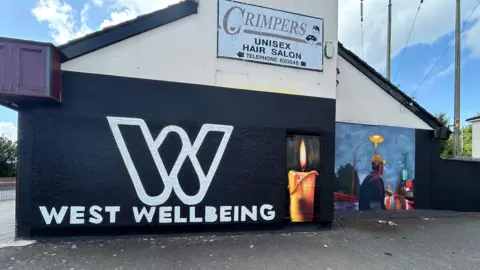 The side of a gable wall is painted black with white lettering stating, West Wellbeing. A painting of a young man is on the next wall.