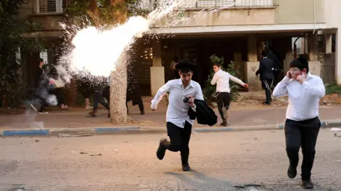 Three ultra-Orthodox Jewish children run from a stun grenade explosion, one covers his ears. The grenade has exploded into a small ball of light a short distance from the young boys.