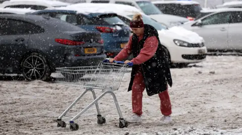 A woman pushes a trolley full of her supermarket shopping through far-stretching, thick brown slush in a car park. She wears a black sleeveless coat and red tracksuit. Her trolley is empty. In the background snow covered cars are visible 