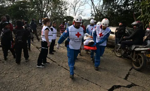 Joaquin Sarmiento / AFP via Getty Images Emergency workers in white vests carry a stretcher with an injured person lying on it, in Cauca on Saturday.